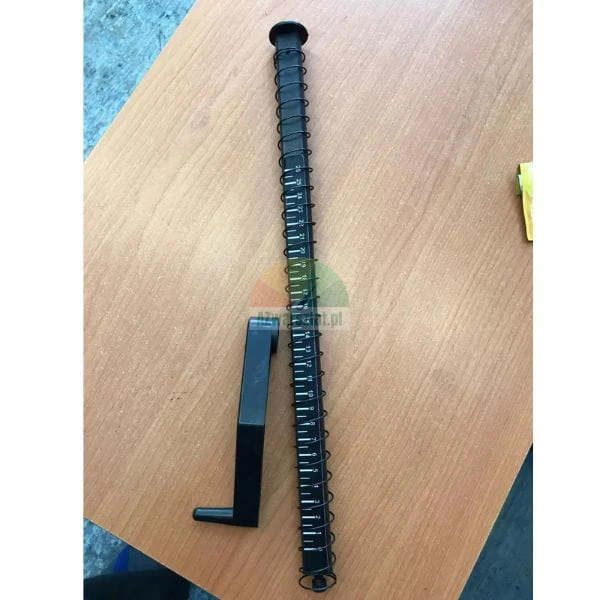 Długi czarny element maszyny do bindowania z podziałką i plastikowa korba na stole.